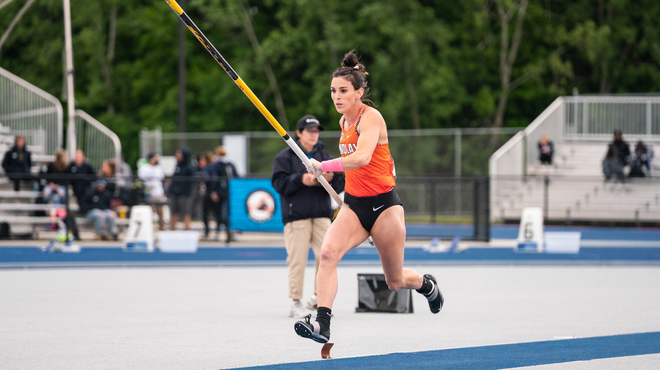 Savannah Amato in orange jersey top and black bottoms running with pole for pole vault