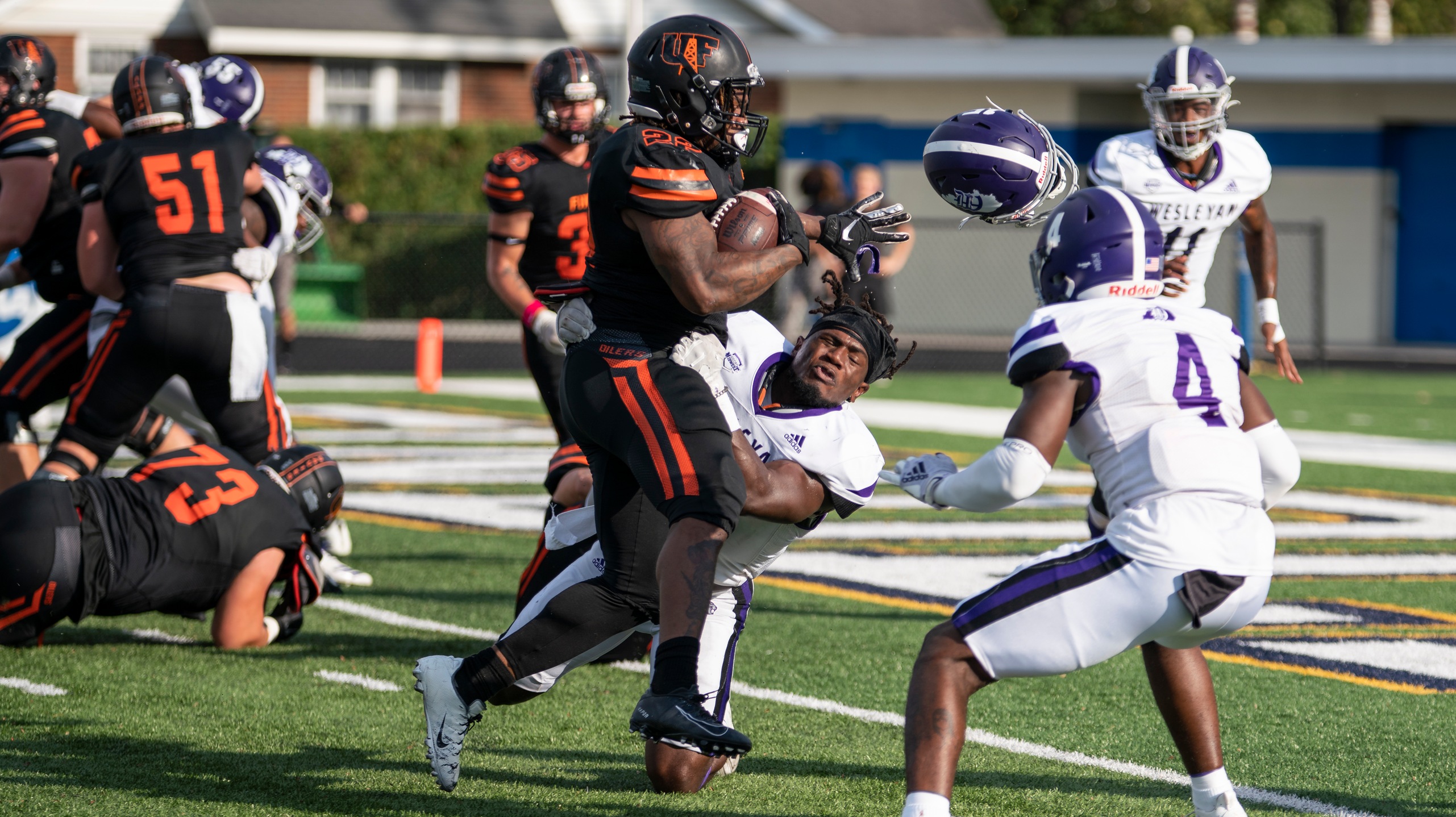 Runner in black jersey running over defender in white with his purple helmet flying off due to forceable contact